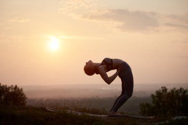 Güneş doğarken çimenli tepeye parlak pastel pembe puslu gökyüzü uzak ufuk yukarıda kopya alan arka plan açık havada bükme jimnastik egzersizleri yapıyor çekici genç kadın silüeti.