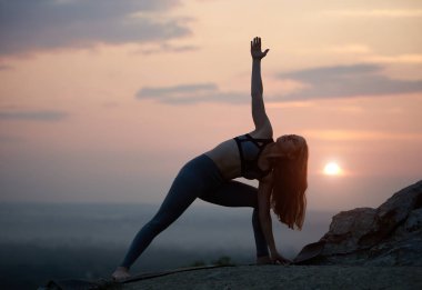 Açık havada bulutlu güzel pembe gökyüzü kopya alan arka plan üzerinde yüksek, kayalık tepe üzerinde bükme yoga egzersizleri yapıyor ve ufuk güneş üzerinde çekici uzun saçlı genç jimnastikçi kadın silüeti.