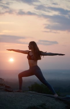 İnce çekici uzun saçlı kız ayakta Yoga gergin kollu silüeti günbatımında gökyüzünün bulutlu güzel sisli pembe arka plan üzerinde büyük kayanın üzerinde açık havada poz. Virabhadrasana