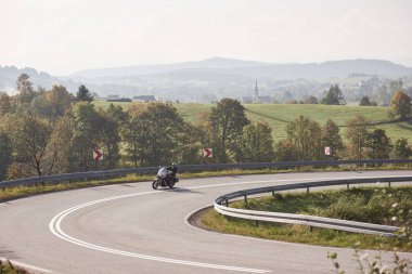 Motosiklet kask ve siyah deri giyim binicilik bisiklet keskin boyunca parlak yaz günü kırsal manzara ve uzak yeşil tepeler puslu arka plan boş yol açmak.