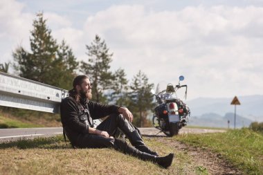 Boyunda atletik kas motosikletçi modern güçlü cruiser motosiklet parlak mavi gökyüzü altında güneşli uzak tepelerin arka plan bulanık çimenli kenarında oturan siyah deri giyim.