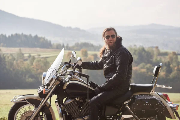黒革のジャケットと緑ののどかな農村風景と光霧空の背景をぼかしの巡洋艦のバイクに座っているサングラスに長髪のハンサムなひげを生やしたモーターサイク リスト — ストック写真