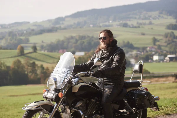 黒革のジャケットと緑ののどかな農村風景の背景をぼかした写真を現代のバイクに座っているサングラスに長髪のハンサムなひげを生やしたモーターサイク リストの側面図 — ストック写真