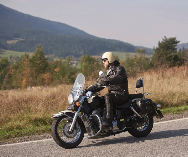 Bearded biker in helmet, sunglasses and black leather clothing riding cruiser bike along sharp turn of empty road on bright summer day on misty background of rural landscape and distant green hills.