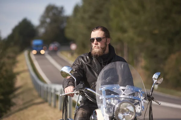 Portrait of handsome bearded biker in black leather jacket and sunglasses sitting on modern motorcycle on roadside on blurred background of twisty road with moving vehicles and green trees.