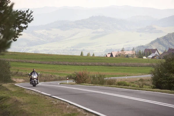 黒革の衣装をツイスト道に沿っての巡洋艦のバイクに乗って青い朝の空の下 遠いで緑の木質の霧丘の背景に明るい日当たりの良い夏の日に単独でモーターサイク リスト — ストック写真