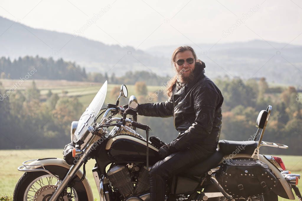Handsome bearded motorcyclist with long hair in black leather jacket and sunglasses sitting on cruiser motorcycle on blurred background of green peaceful rural landscape and light foggy sky.