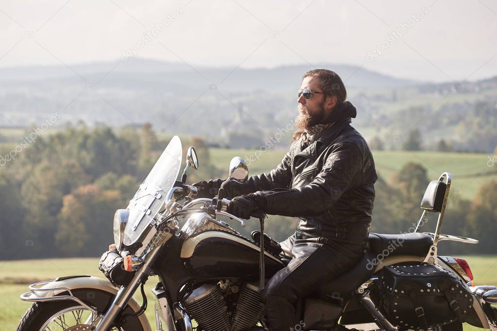 Side view of handsome bearded biker with long hair in black leather jacket and sunglasses sitting on cruiser motorcycle on blurred background of green peaceful rural landscape and light foggy sky.