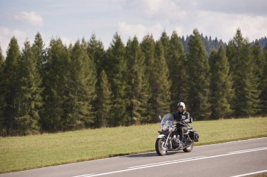 Kask, güneş gözlüğü ve yüksek yoğun çam ağaçları orman ve açık parlak mavi gökyüzü arka plan üzerinde siyah deri giyim binicilik bisiklet gün güneşli yaz boş asfalt yolda sakallı motosikletçi.