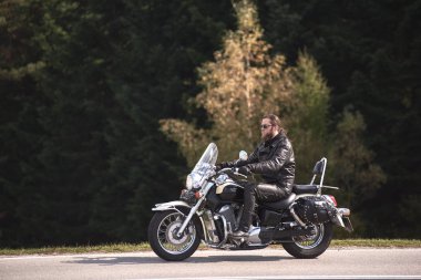 Güneş gözlüğü ve güneşli autumnr gün orman arka plan üzerinde dar asfalt yol boyunca Kruvazör motosiklet sürme siyah deri giyim uzun saçlı sakallı motosikletçi yan görünüm.
