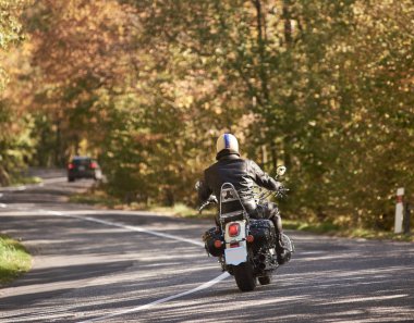Motosikletçi siyah deri ceket ve yeşil orman ağaçları bitki örtüsü ve hareketli araba bulanık arka plan üzerinde parlak güneşli yaz gününde bükülmüş asfalt yol boyunca motosiklet sürme kask arkadan görünüm.