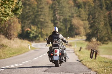 Arkadan görünüşü motorcu siyah deri ceket ve motosiklet virajlı yol boyunca parlak güneşli yaz gününde yoğun yeşil orman ağaçları bulanık arka plan üzerinde binme kask. Aktif yaşam tarzı ve seyahat.