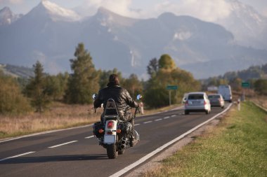 Araçlar ve mavi gökyüzü parlak güneşli gün hareketli motosikletçi karlı zirveleri ile güzel dağ aralığının arka plan bulanık yol boyunca motosiklet sürme siyah deri ceketli arkadan görünüm.