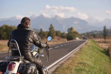 Motorcu Kruvazör motosiklet yol boyunca karlı zirveleri ve mavi gökyüzü ile güzel dağ aralığının arka plan bulanık parlak güneşli sonbahar gün sürme siyah deri ceketli arkadan görünüm.