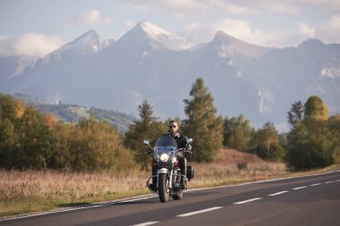 Güneş gözlüğü ve siyah deri giyim beyaz karlı zirveleri ve parlak sonbahar gökyüzü kopya alan arka plan mavi sisli dağlar üzerinde modern güçlü cruiser motosiklet sürme sakallı motorcu.
