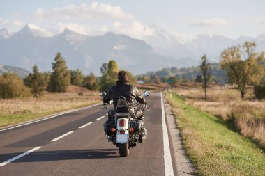 Araçlar ve mavi gökyüzü parlak güneşli yaz günü hareketli motorcu karlı zirveleri ile güzel dağ aralığının arka plan bulanık yol boyunca motosiklet sürme siyah deri ceketli arkadan görünüm.