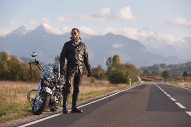 Karanlık güneş gözlüğü, siyah deri giyim daimi yeşil kırsal manzara, sarp dağ zirveleri ve parlak mavi gökyüzü arka plan üzerinde modern güçlü motosiklet, boyunda atletik motorcu sakallı.