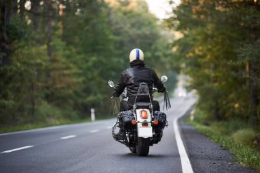 Arkadan görünüşü motorcu siyah deri ceket ve beyaz kask uzun yeşil ağaçlar arasında engebeli yolda bir motosiklet sürme içinde. Aktif yaşam tarzı, aşk maceraları kavramı için.