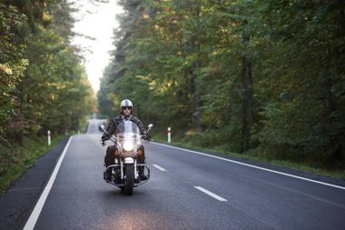 Güneş gözlüklü, kasklı, siyah deri elbiseli, asfalt yolda motosiklet süren sakallı motorcu güneşli yaz akşamlarında uzun yeşil ağaçların arasında dolanıyor..