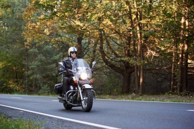 Güneş gözlüğü, kask ve siyah deri giyim binme Kruvazör yakışıklı sakallı motorcu motosiklet uzun yeşil ağaçlar arasında güneşli yaz akşamı sarma asfalt yol boyunca
