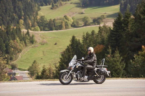 ヘルメット サングラス 黒革の衣類が狭いアスファルトの道に沿ってクルーザー バイクに乗って背の高い木々 やグリー草の背景の日当たりの良い夏の日にひげを生やしたモーターサイク リストの側面図 — ストック写真