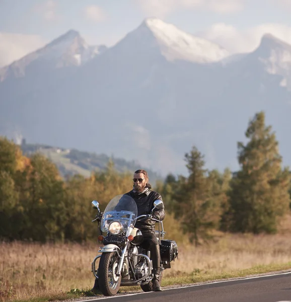 黒革のジャケットと緑の田園風景 遠く白い日当たりの良い山の峰の背景をぼかしの国沿道の巡洋艦のバイクに座っているサングラスに髭があるバイク — ストック写真