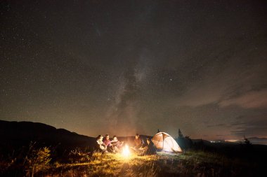Beş mutlu turistlere yolcular gitar, erkek ve yakınında çadır, kamp ateşi yakarak oturan kadın güzel kamp gece zevk ile şirketin 