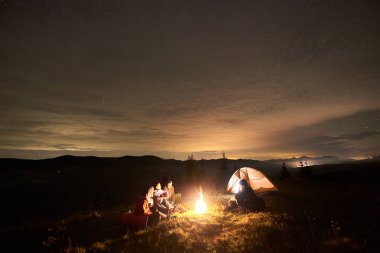 Beş mutlu turistlere yolcular gitar, erkek ve yakınında çadır, kamp ateşi yakarak oturan kadın güzel kamp gece zevk ile şirketin 
