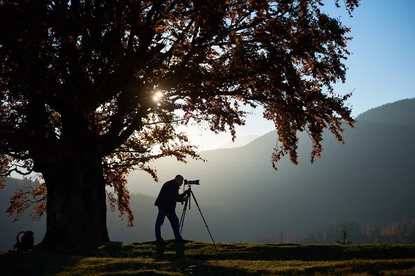 Silhouette of tourist photographer using tripod and professional camera and taking picture of mountains panorama on sunset background