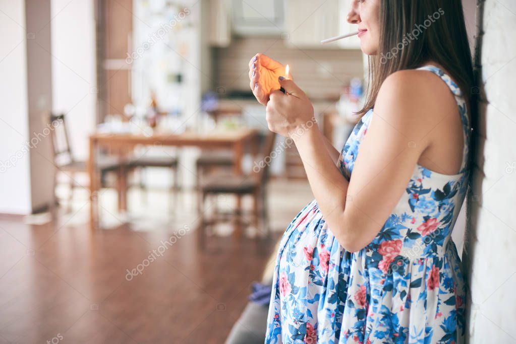 Cropped view of pregnant woman in dress standing in kitchen and keeping in hands cigarette and lighter