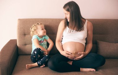 Sevimli kız evde kanepe, gülümseyen hamile anne olarak bakıyor ve gülüyor