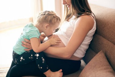 Kanepe ve sevimli kızı öpüşme ve büyük göbek kucaklayan üzerinde oturan mutlu hamile anne yan görünüm