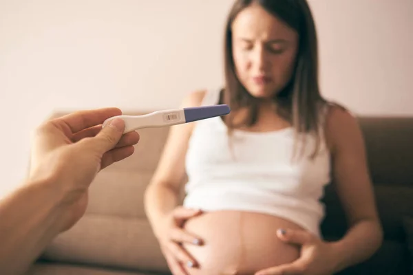 Selective focus of pregnancy test in male hand and disappointed young woman sitting on sofa and keeping belly with hands