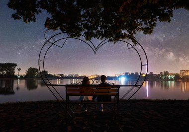 Rear view of man and woman sitting on bench near campfire on lake shore \