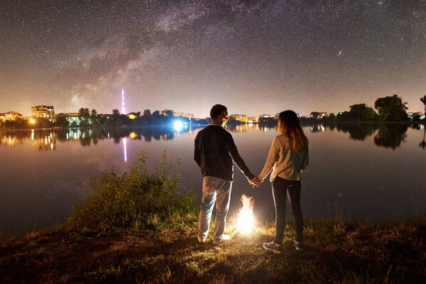Rear view of man and woman standing near bonfire at night camping on lake shore 