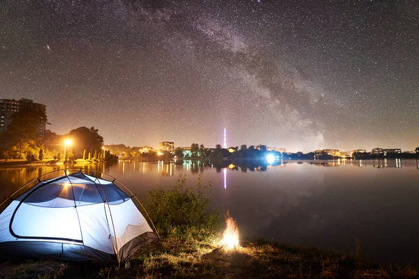 Illuminated tent and campfire on lake shore under starry sky