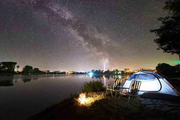 Illuminated tent and campfire on lake shore under starry sky