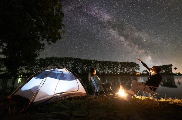 back view of man and woman friends sitting on chairs near campfire and illuminated tent and enjoying sky full of stars and Milky way