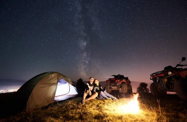 Happy couple man and woman trevelers sitting near tourist tent and burning campfire