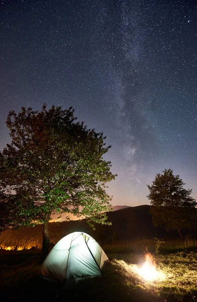 Illuminated tent and campfire under magical night sky full of stars and Milky way