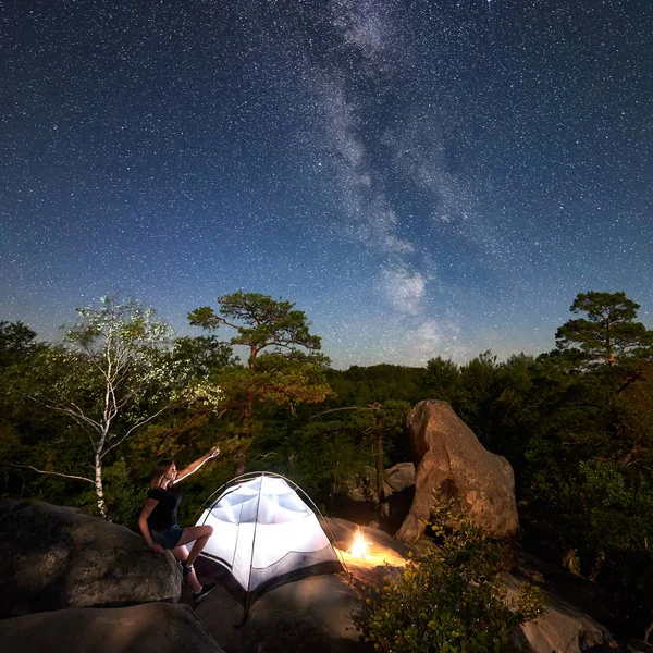 Young woman hiker having rest on rocky mountain top beside camp, campfire and glowing tourist tent at summer nighttime