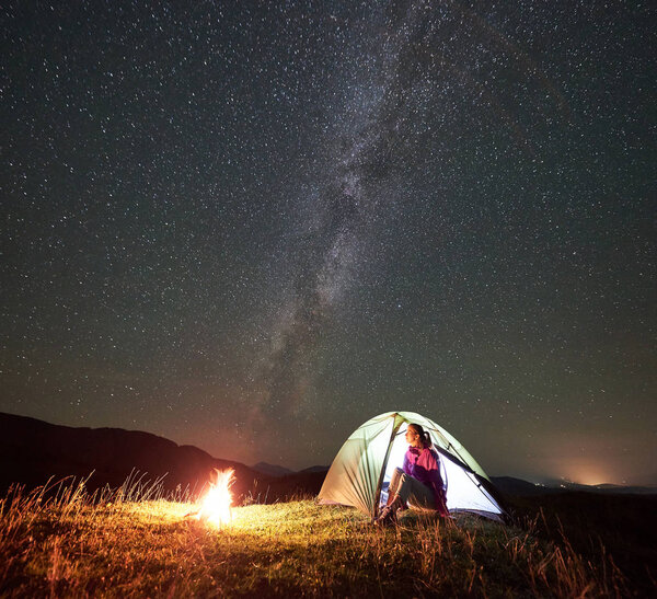 Female hiker resting at summer night camping in mountains near campfire 
