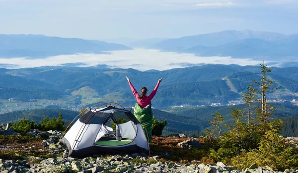 Happy hiker woman in pink sweater and sleeping bag resting beside tourist tent