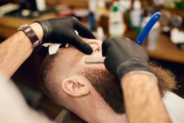 Müşteri sakallı yüzünde keskin bıçak ile eldiven berber elleri ile tıraş için sandalyede uzanmış