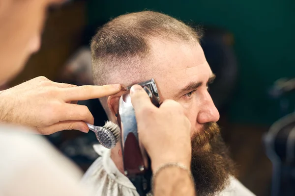 Young barber working with electric razor making haircut for stylish bearded man sitting in hairdresser chair