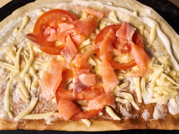 top view of sliced salmon fish and tomato, grated cheese on round thin and baked dough base greased with white sauce