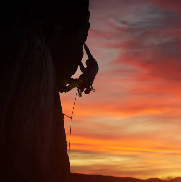 Side view of male silhouette climbing on natural rocky wall on orange sunset over mountains background