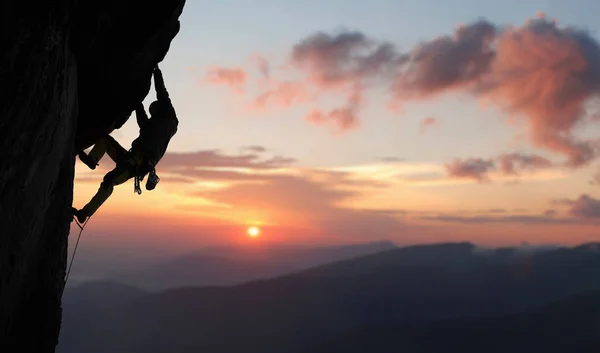 在日落征服陡峭悬崖顶端的岩石墙上 男性登山者的低角度视图 — 图库照片