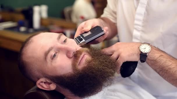 美容椅子に座っている男性のひげの口ひげクライアントのための電気かみそりマシンで顔を剃る巧みな手 — ストック動画