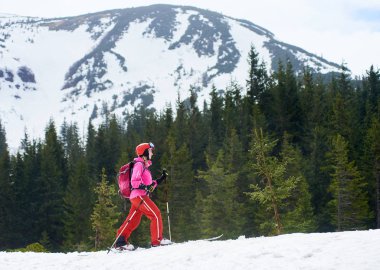 Pembe takım elbiseli, sırt çantalı, karlı dağ eteklerini keşfeden ve kayak yapan genç bir bayan kayakçının yan görüntüsü.